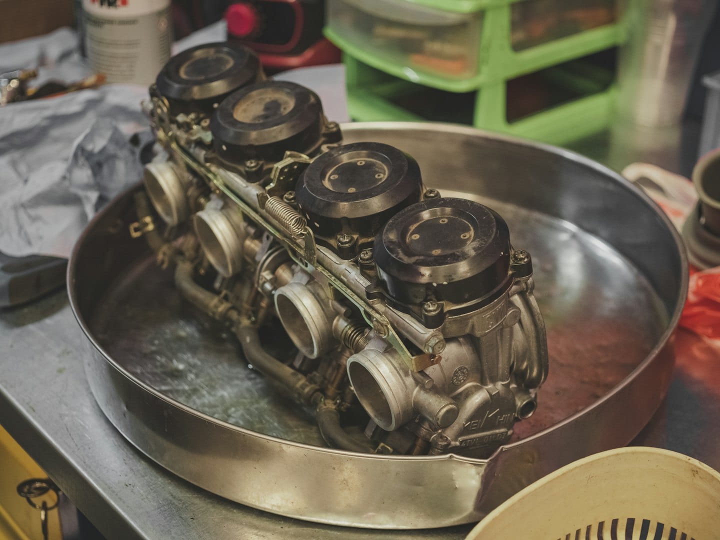 Dört motosiklet karbüratörü bir tezgah üzerindeki metal bir leğende duruyor.